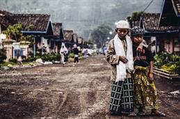 Two Little indonesian Boys are Standing on a muddy street. The taler one has put his arm around the other, they are looking into the camera. In the Background you can see small houses, Little front yards.